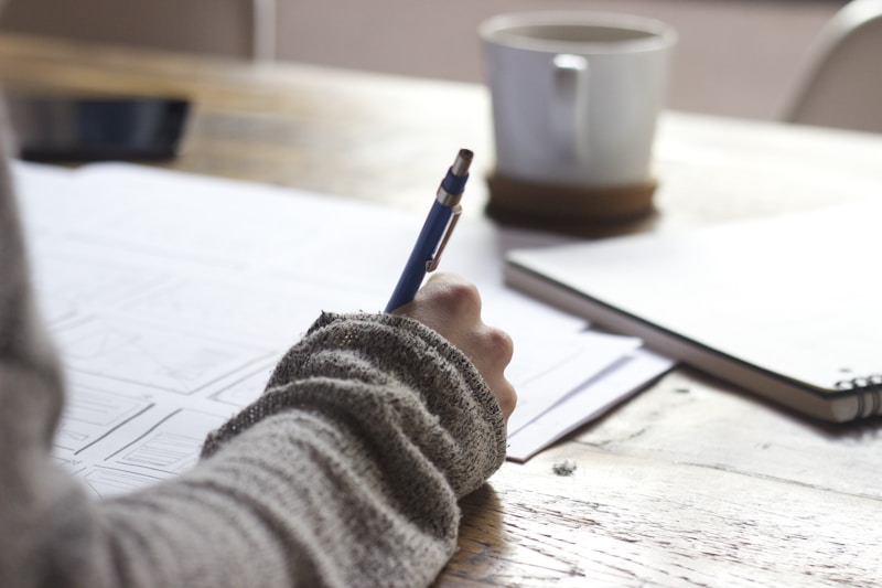 Close-up of a hand writing on a paper grid layout with a blue pen on a wooden table beside a white mug and spiral notebook