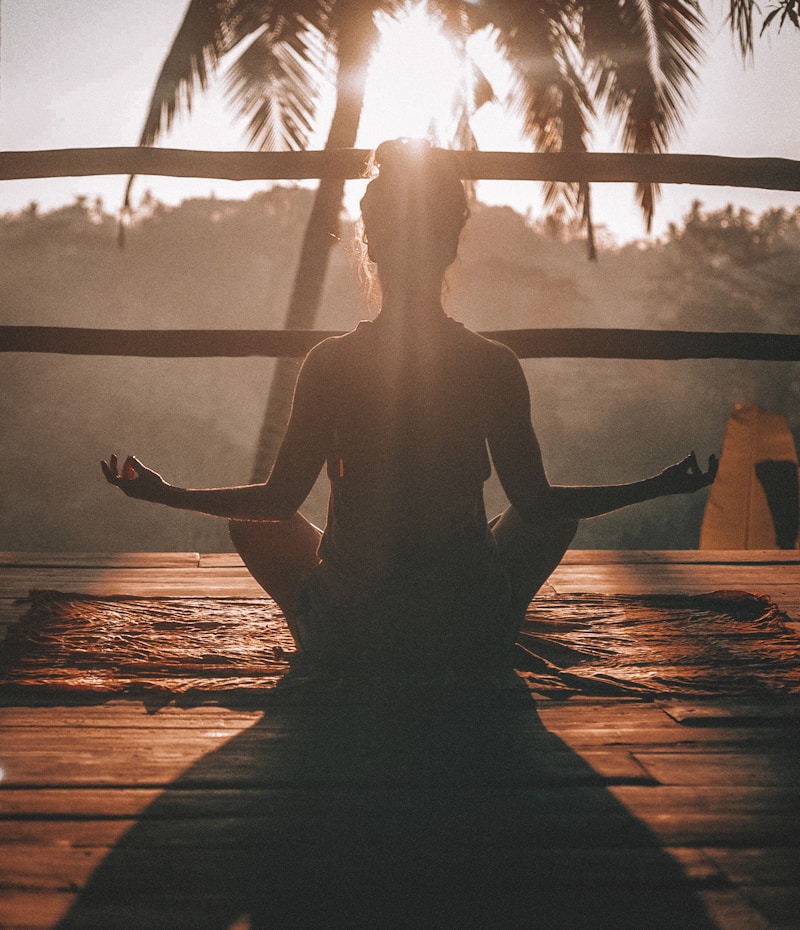 Silhouette of a person meditating in cross-legged pose on a wooden deck at sunrise with palm trees behind a rustic railing